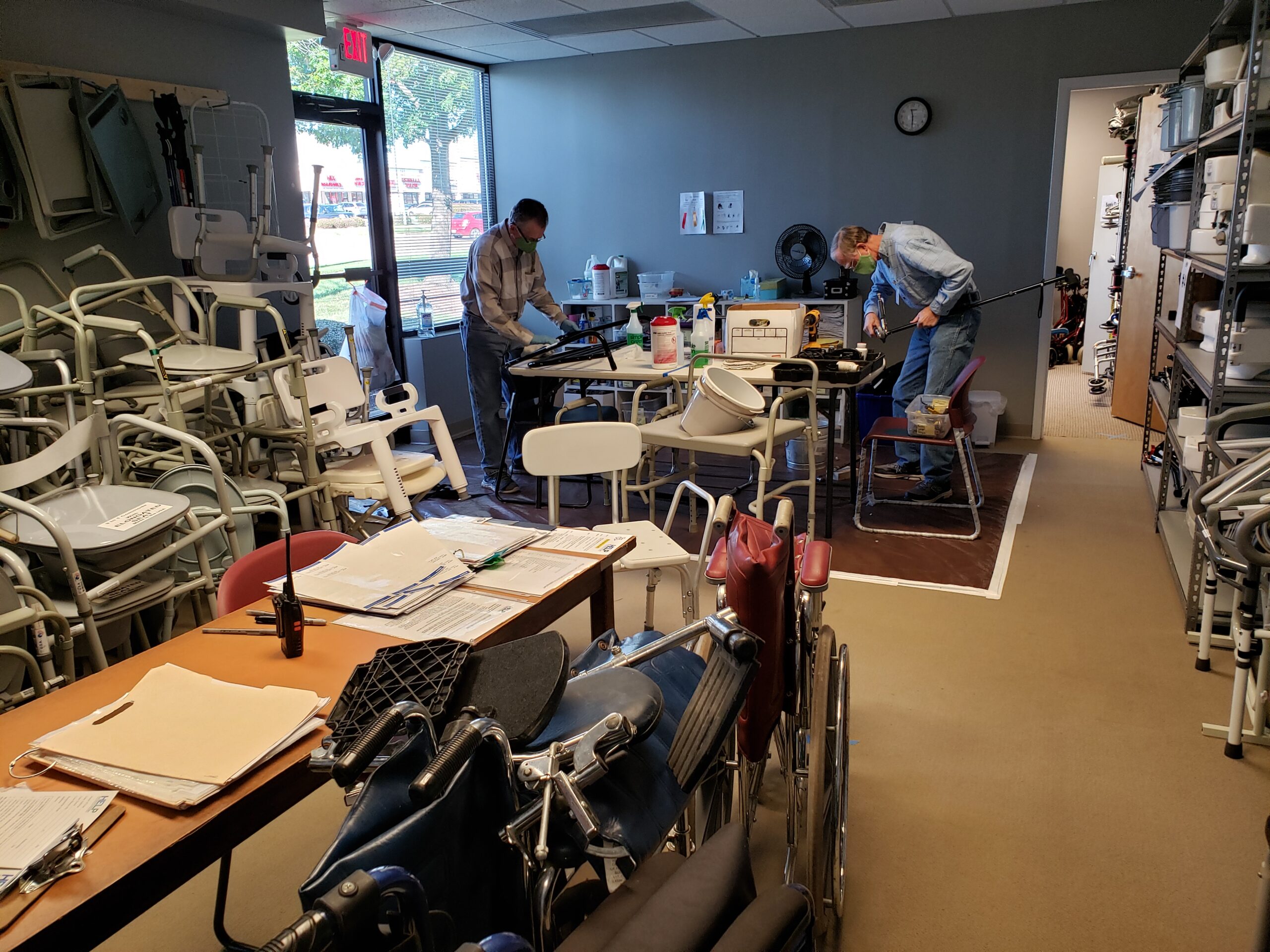 HELP office with piles of different medical equipment around and two volunteers repairing and cleaning items on a table in the back. 